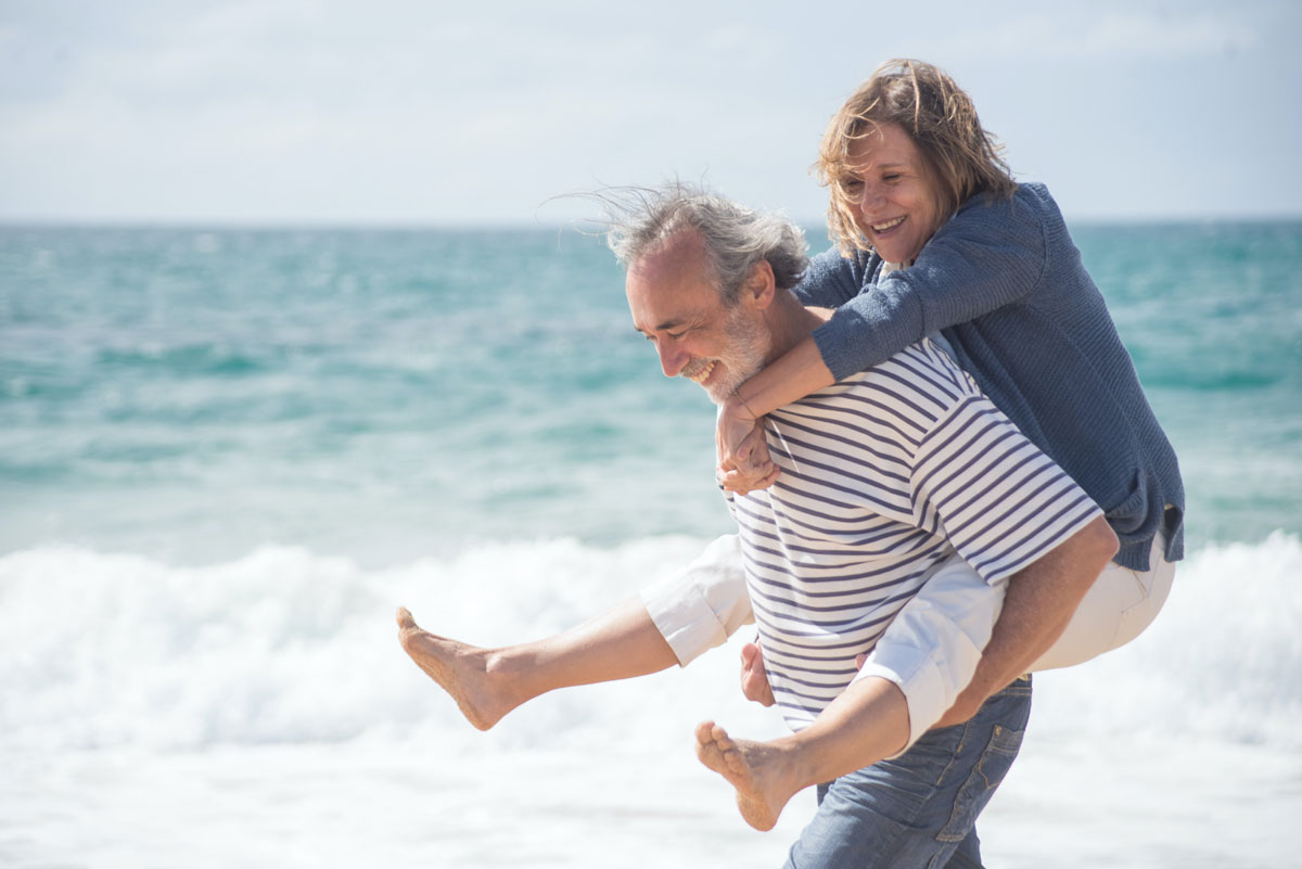 Smiling older couple on beach, man is carrying woman piggyback.