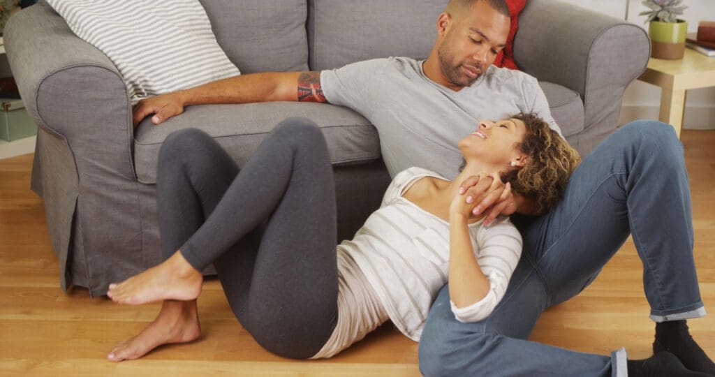 A relaxed couple sitting on the floor in front of a couch, holding hands and smiling at each other lovingly in a cozy living room.